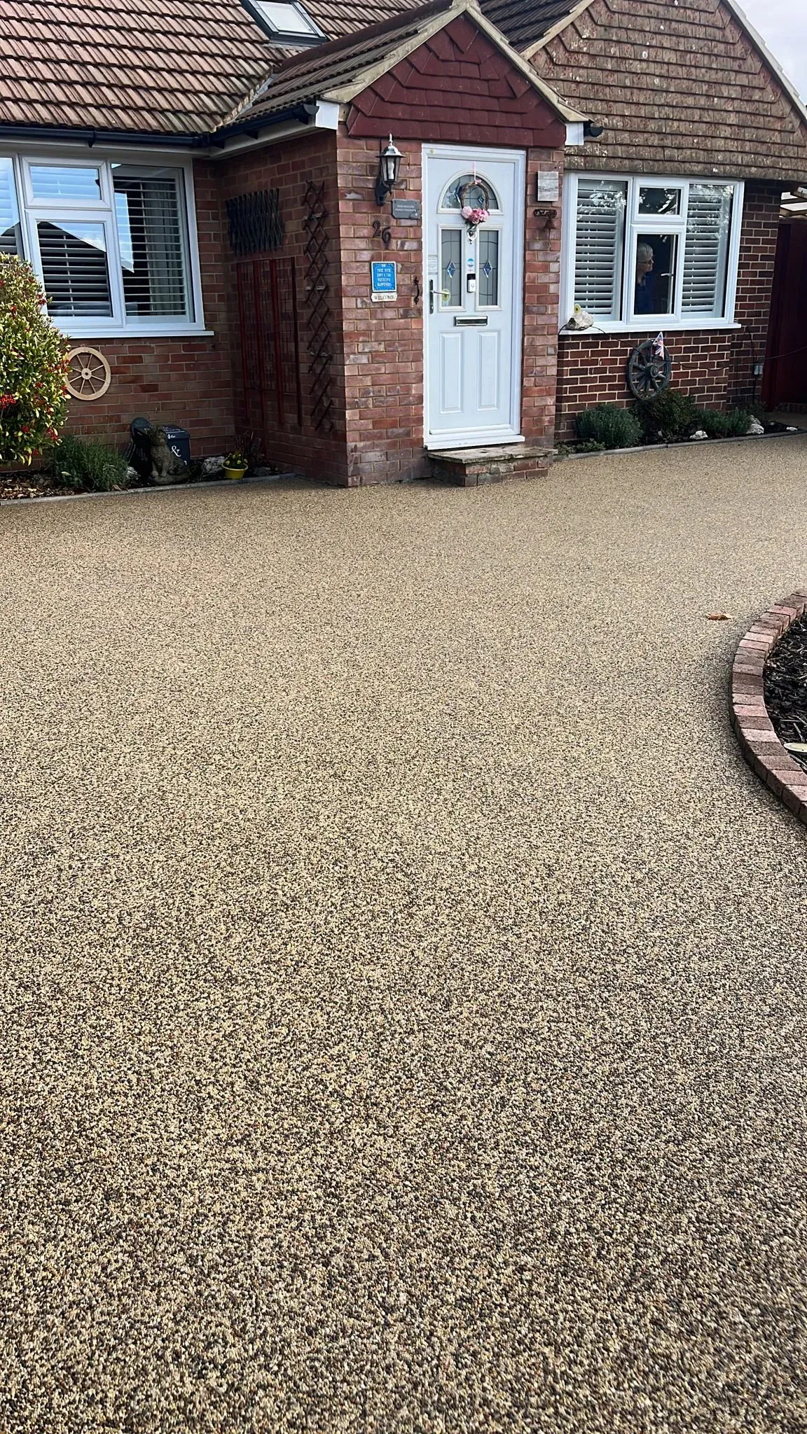 A full view of a modern resin-bound driveway in a warm beige tone, leading to the entrance of a brick-built house with a white front door, demonstrating a complete driveway transformation.
