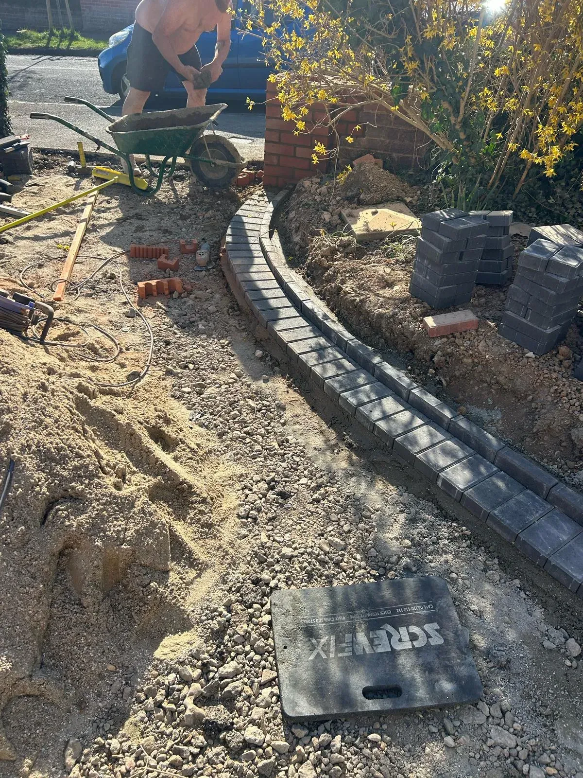 Construction site for a new driveway or garden path, with a worker in the background and a curved border of dark grey block paving being laid.