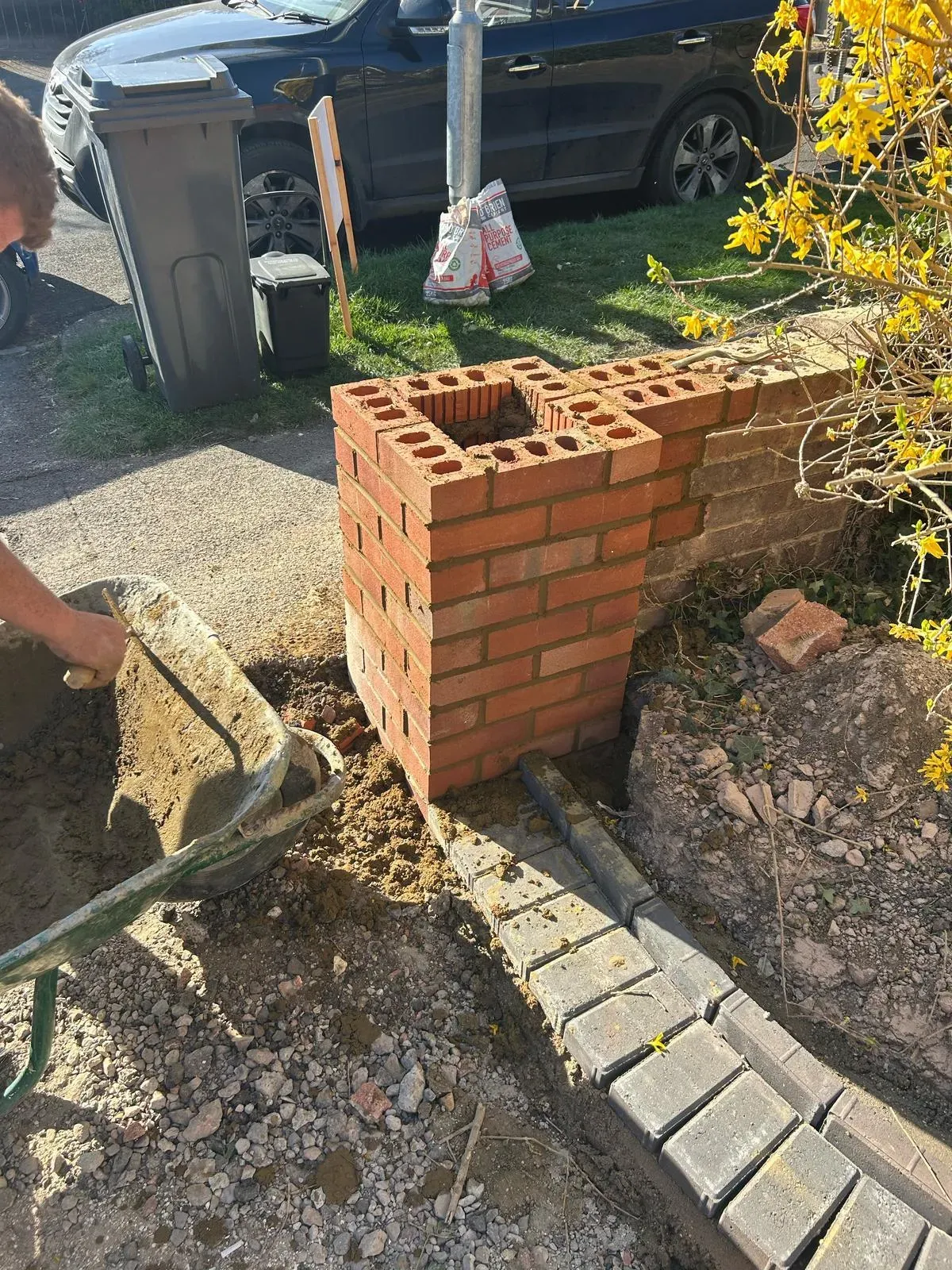 Work in progress showing a new brick pillar being built, block paving being laid for an edge or path, and a wheelbarrow with soil during groundwork.