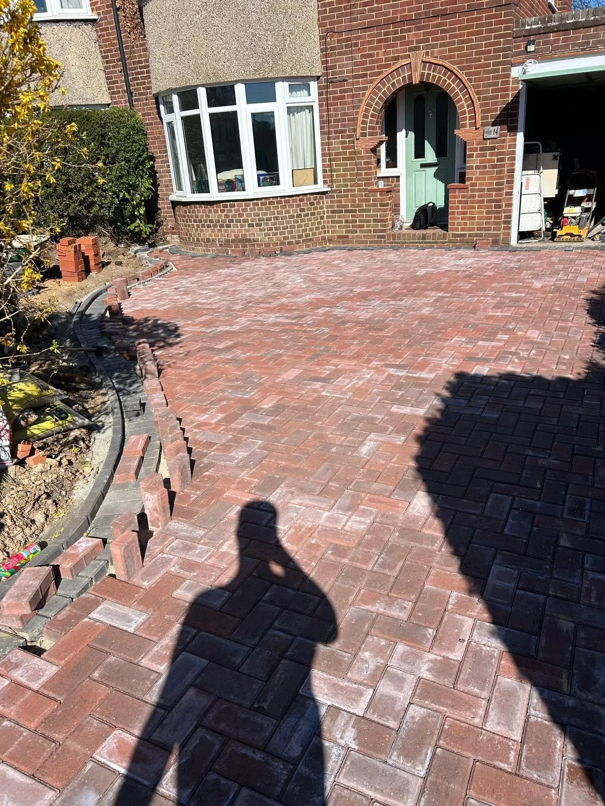 Block paving driveway installation in progress, showing red and brown pavers laid in a herringbone pattern, with an unfinished border and stacks of bricks on the left.