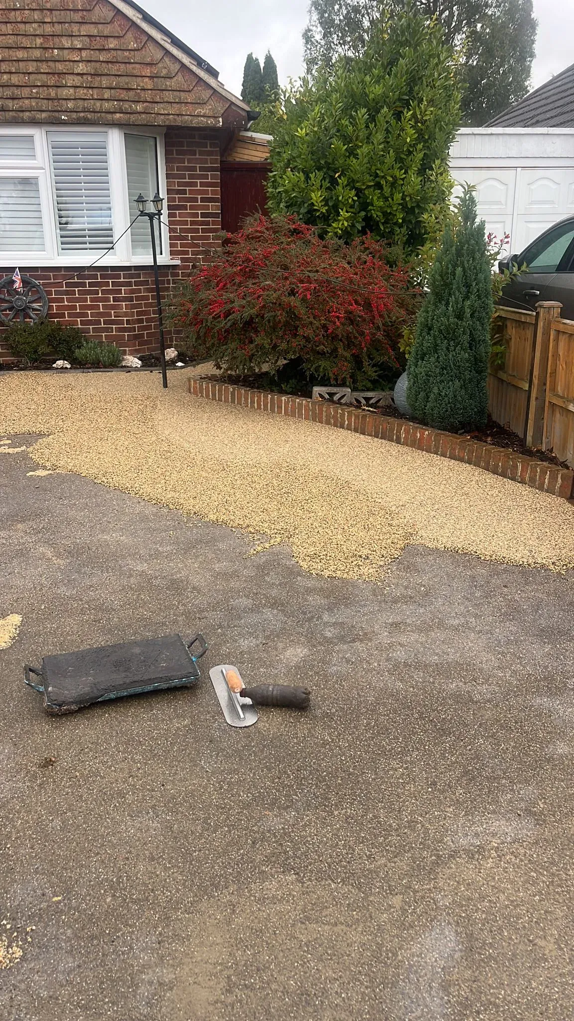 Construction scene of a resin-bound driveway installation, showing a fresh layer of light-coloured aggregate being smoothed over an old tarmac driveway in front of a brick house with garden shrubs. A trowel and float lie on the ground.