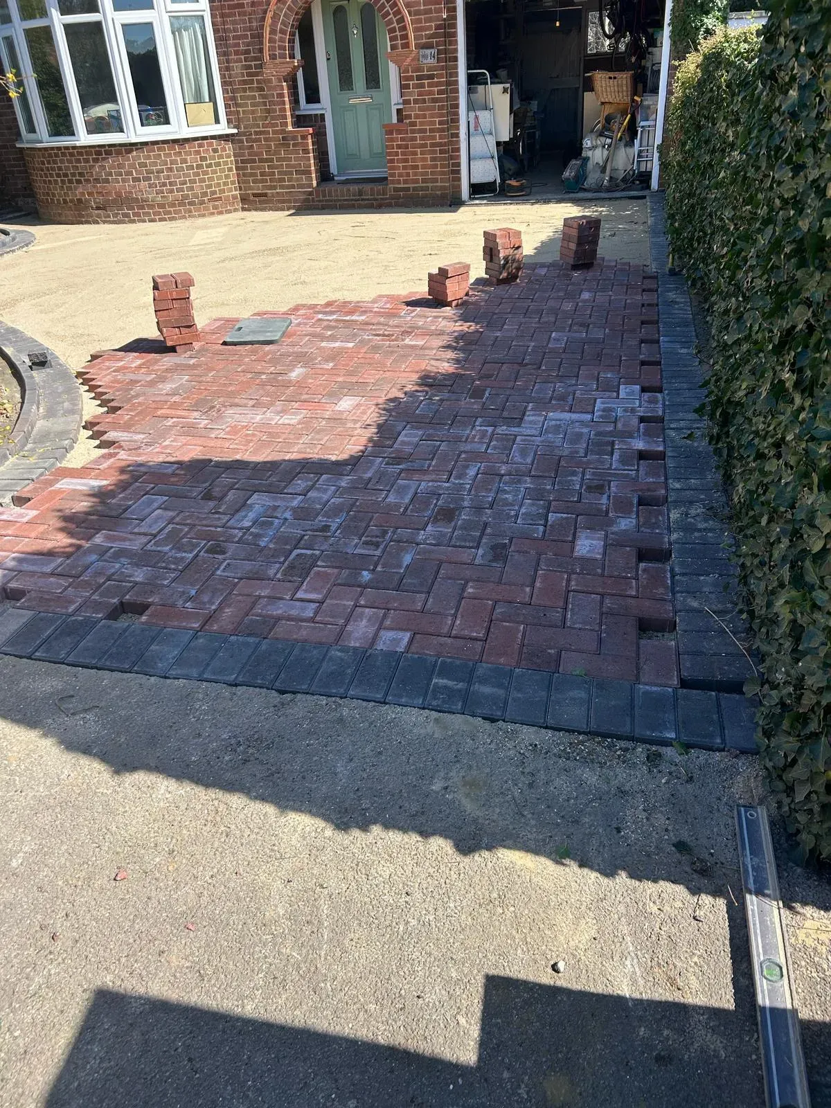 A partially completed block paving driveway, with red pavers and a dark border laid on a sand sub-base, in front of a house and beside a hedge.