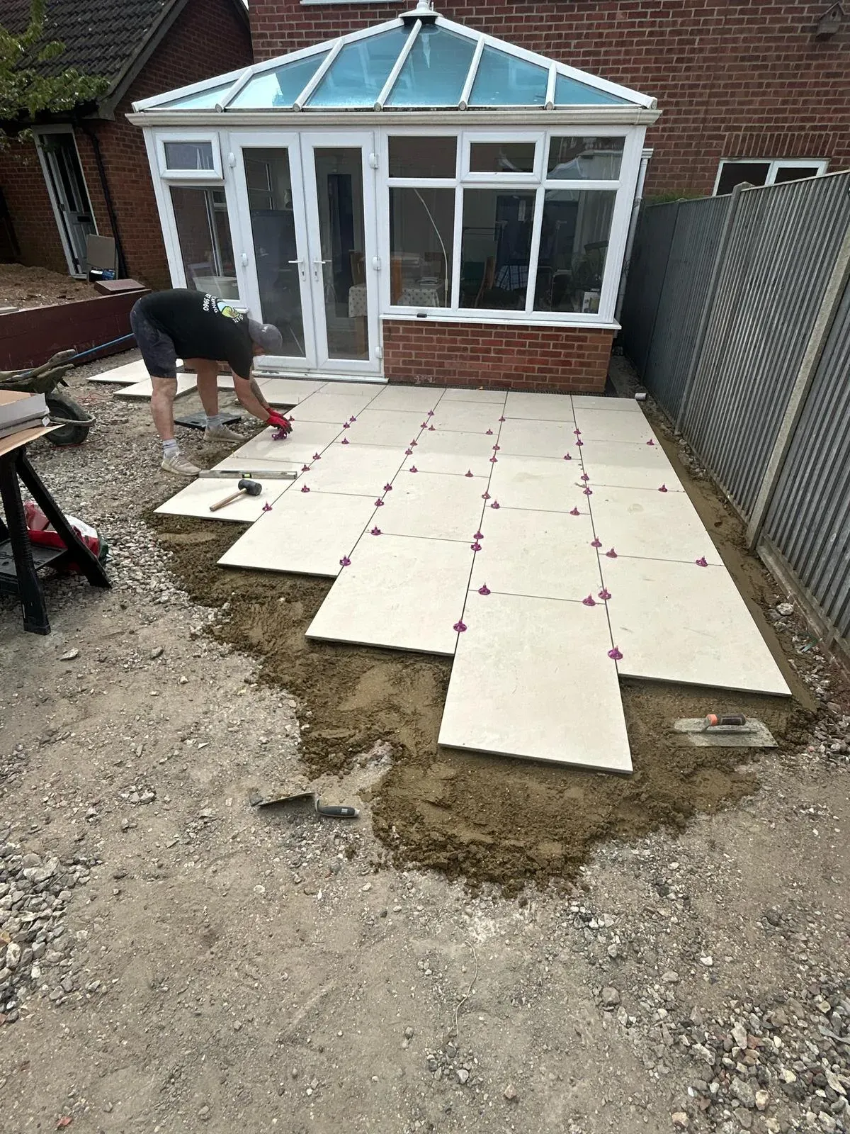 A professional laying light-coloured patio slabs in a garden, with some already laid and spaced with purple clips, next to a white conservatory and brick house.